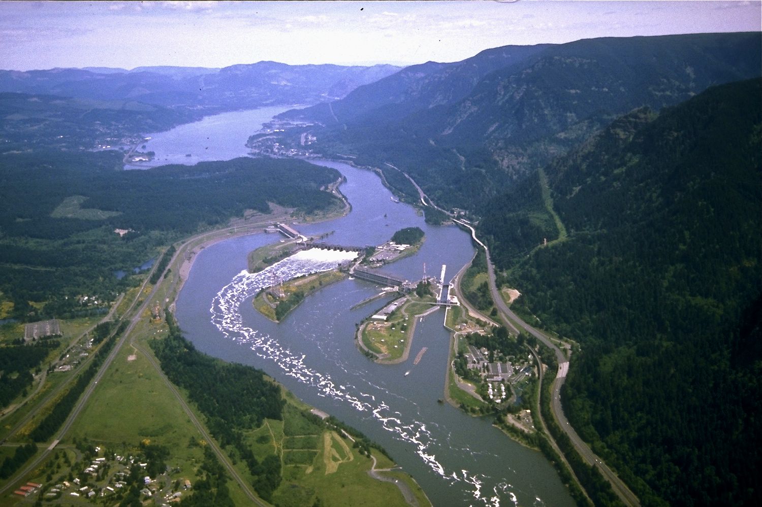 Aerial view looking east up the Columbia River gorge, with Bonneville Dam, its spillway and navigation lock spanning the river.